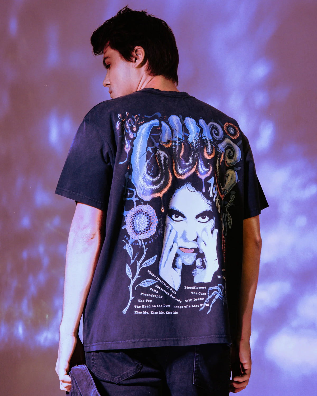 RVCA x The Cure Black Graphic Tee shown on a model under purple lighting, highlighting the oversized psychedelic back artwork and vintage-inspired band design.