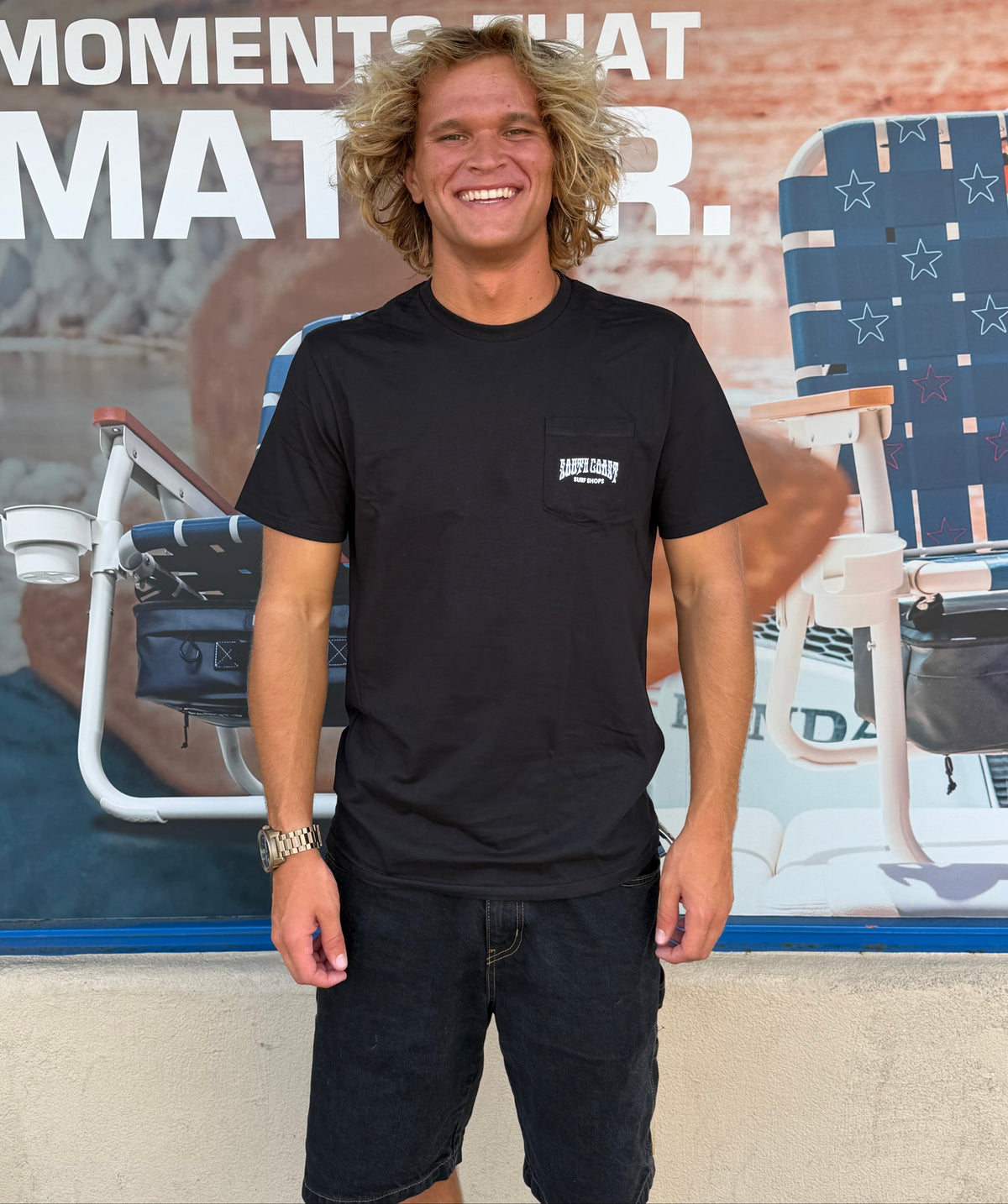 A smiling man with curly blond hair stands outdoors wearing a black short-sleeve pocket T-shirt with a small white chest logo and dark denim shorts, posed in front of a large printed backdrop featuring beach chairs and partial text.