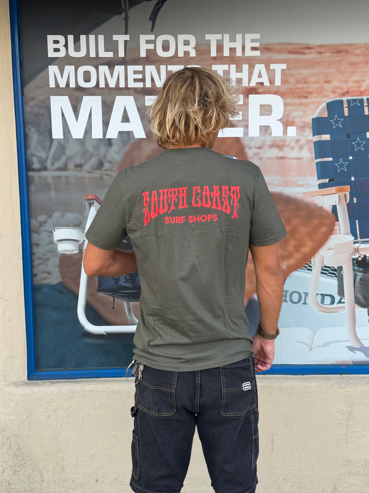 South Coast Drip Tee in Olive featuring a bold red South Coast Surf Shops graphic on the back, modeled from behind.