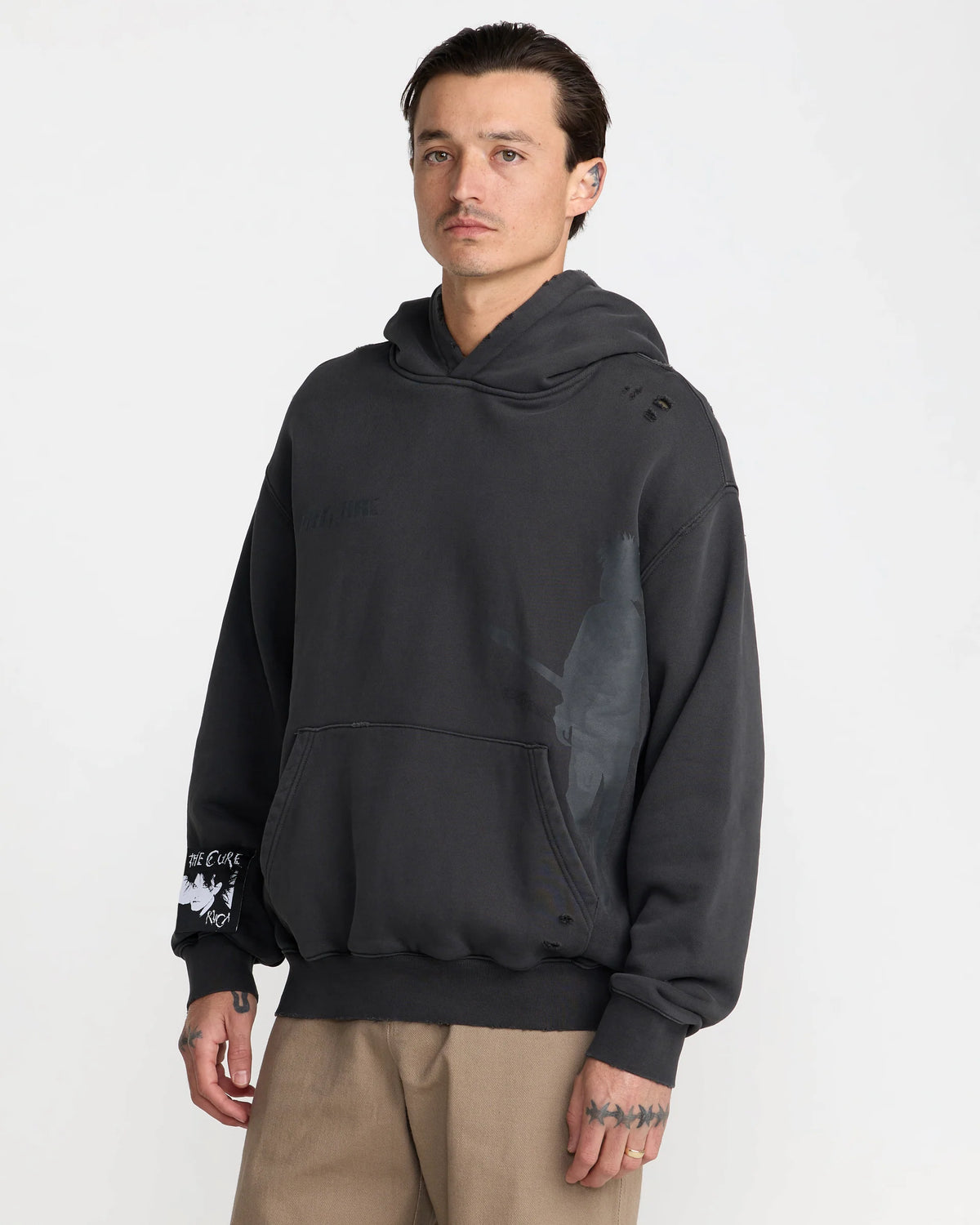 The Cure Washed Black Hoodie shown on a model from an angled front view, highlighting the oversized silhouette, subtle guitarist graphic, and distressed details.