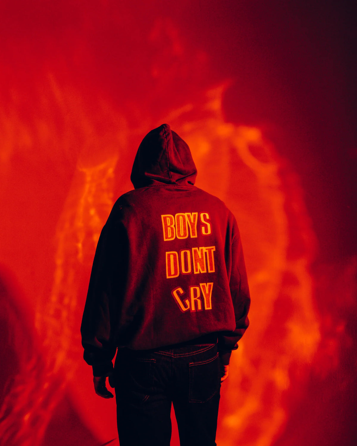 The Cure Boys Don't Cry Hoodie in washed black photographed from behind against a dramatic red background, highlighting the bold back graphic and vintage band aesthetic.